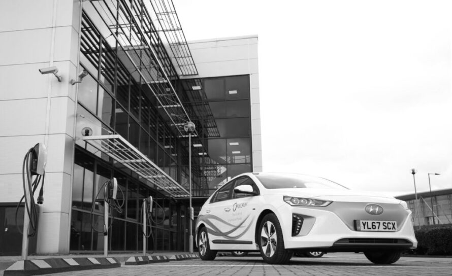 White car with Fulcrum branding on it parked outside a modern building lined with electric chargers available across the front